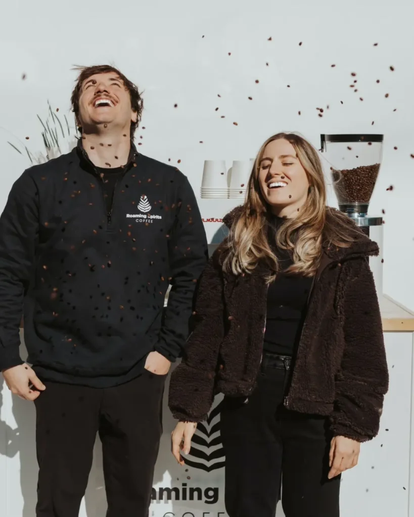 Zack and Bri, founders of Roaming Spirits Coffee, standing and smiling in front of a mobile espresso cart while coffee beans fall from the sky